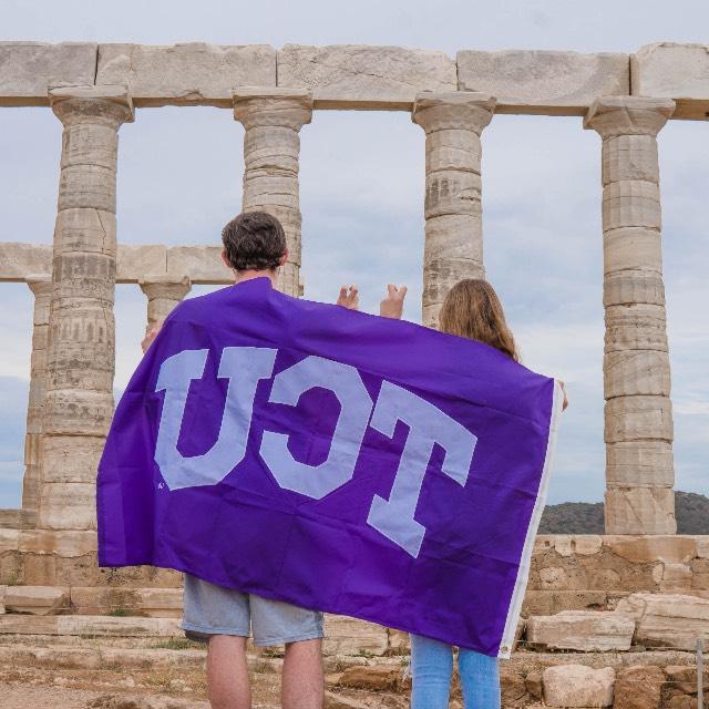 Two students, backs toward us, drape a TCU flag across their shoulders as they look at the pillars of a classical monument